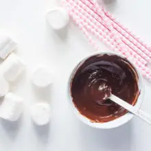 A bowl of melted chocolate sits next to vegan marshmallows and pink paper straws.