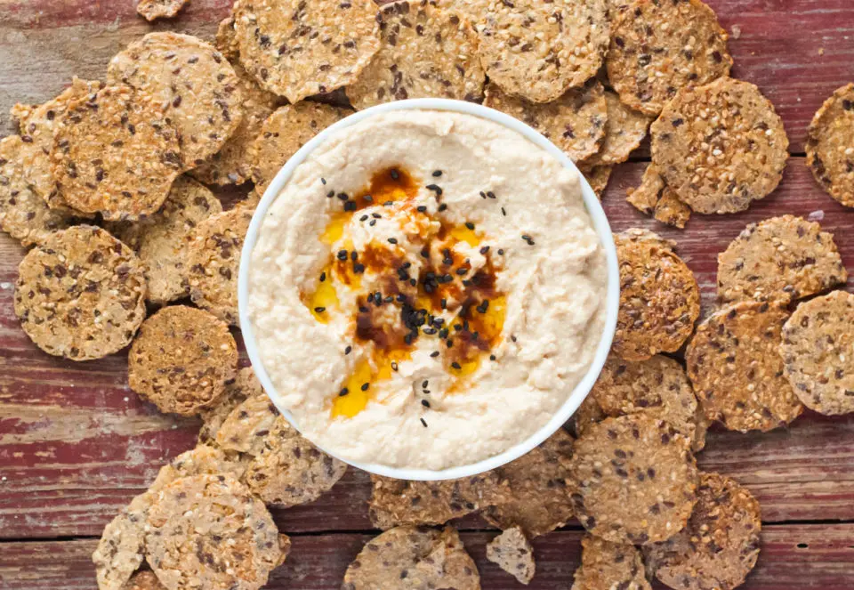 Looking down on a bowl of creamy chickpea dip with black sesame seeds and olive oil over the top. It's surrounded by malt-grain crackers.