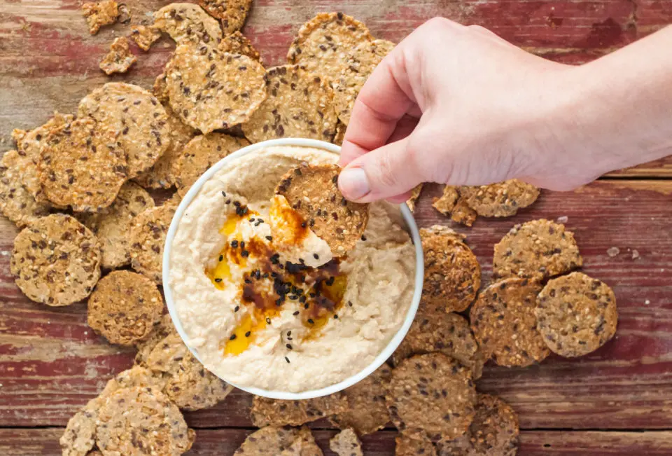 A hand reaches in with a cracker to dip into a bowl of hummus.