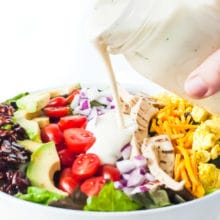 Vegan ranch dressing is poured over a bowl of salad topped with bacon flavored almond slivers, sliced avocados, sliced cherry tomatoes, chopped red onions, vegan cheddar, and tofu scramble.