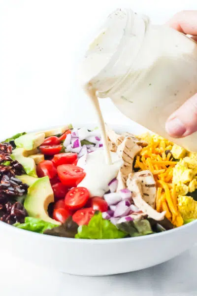 Vegan ranch dressing is poured over a bowl of salad topped with bacon flavored almond slivers, sliced avocados, sliced cherry tomatoes, chopped red onions, vegan cheddar, and tofu scramble.