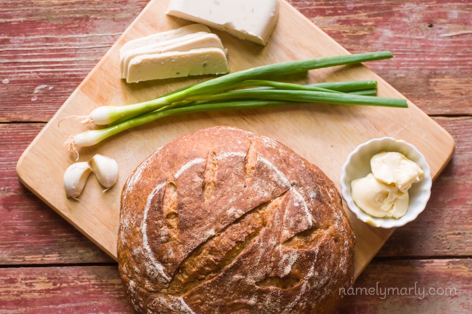 A round loaf sits on a cutting board with vegan butter, green onions, and vegan cheese.