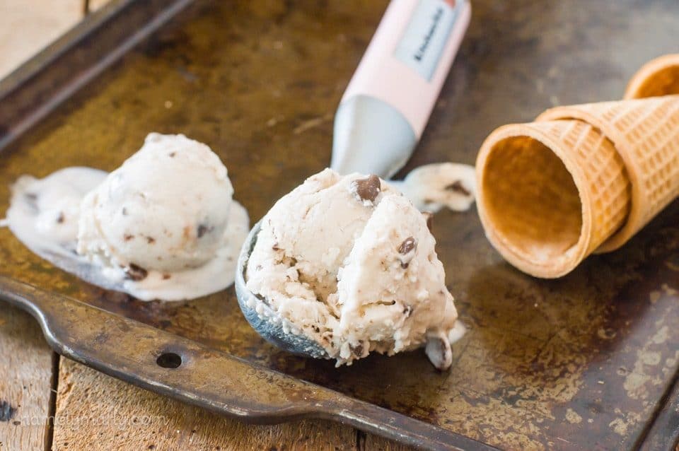 An ice cream scoop holds a scoop of vegan ice cream, with another scoop of ice cream beside it and sugar cones.