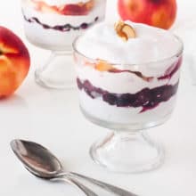 Two serving dishes hold a berry parfait mixture. Two peaches sit beside them.