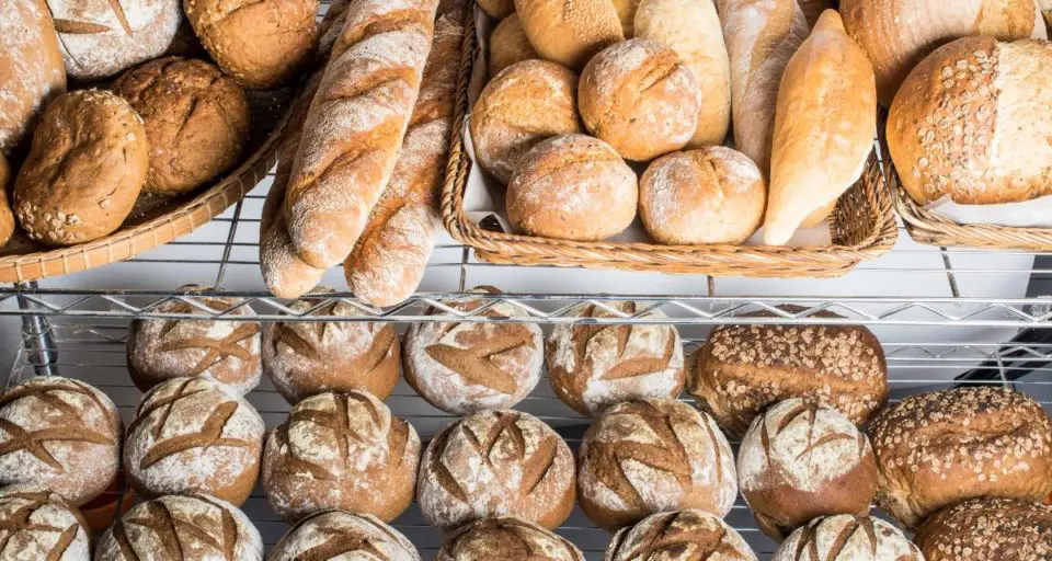 Wire racks in a bakery store hold a variety of different types of bread, including baguettes and artisan round loaves.