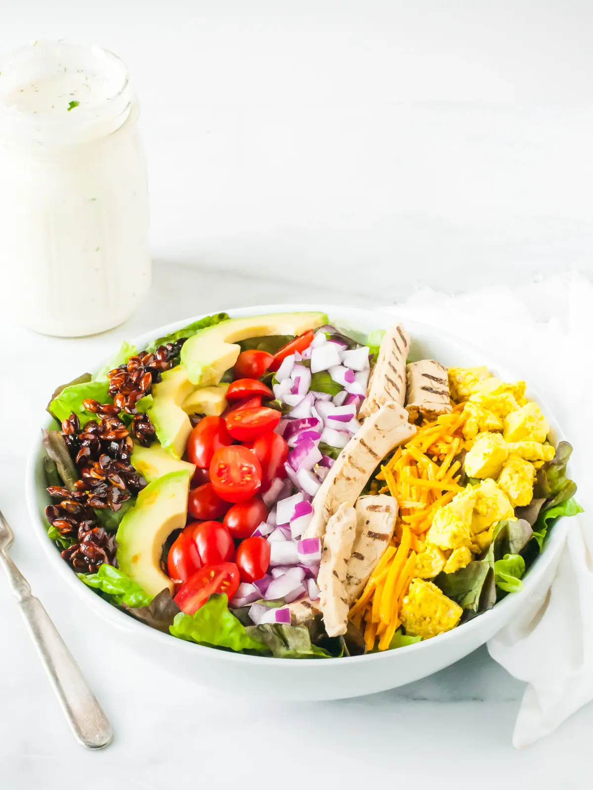 Vegan Cobb Salad in a white bowl with long strips of smoky almond slivers, sliced avocados, vegan cheese, and other toppings. A bottle of vegan ranch dressing is in the background.