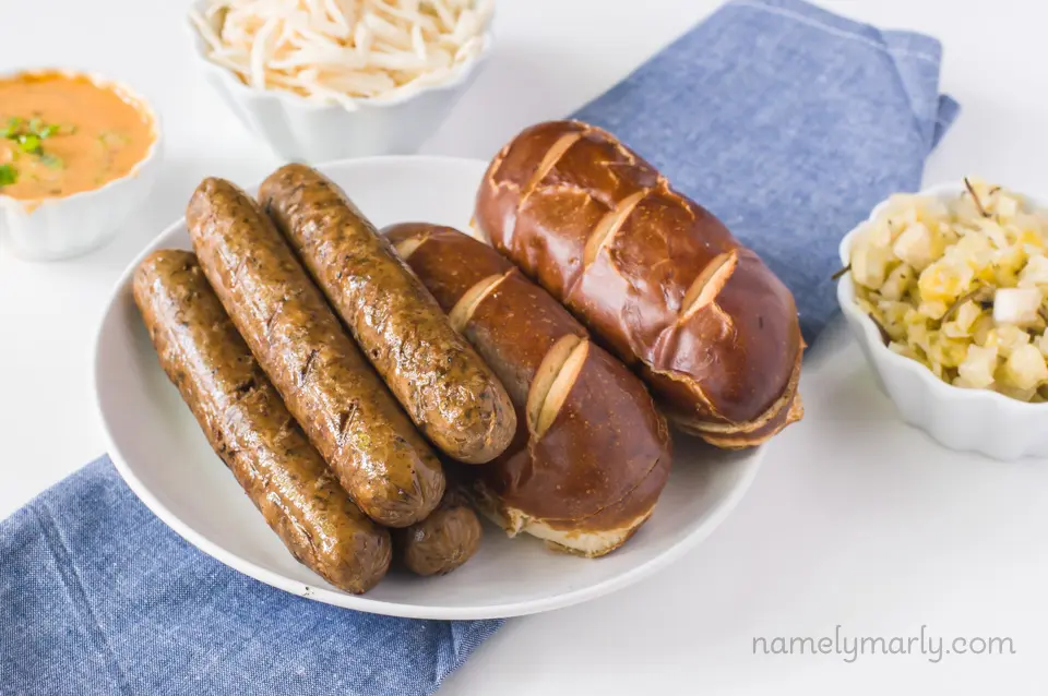 Several vegan brats on a plate next to hot dog buns and surrounded by bowls with other ingredients like vegan mozzarella shreds, and vegan special sauce.