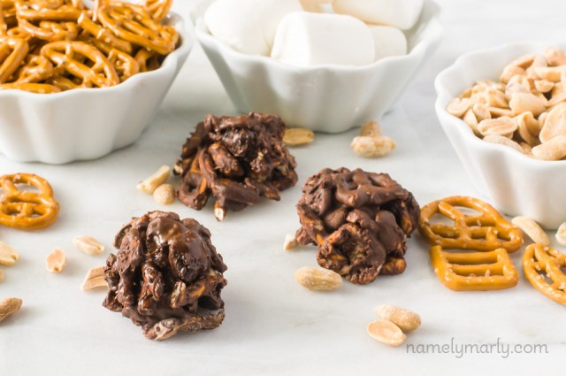 Several chocolate coated cluster cookies are sitting next to bowls of peanuts, vegan marshmallows, and pretzels.