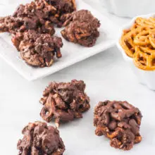 Several Chocolate-Covered Trail Mix Clusters on a white counter top with a bowl of pretzels, vegan marshmallows and more cookies behind them.