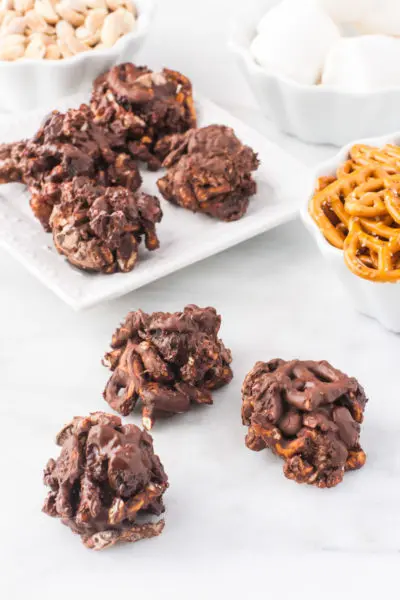 Several Chocolate-Covered Trail Mix Clusters on a white counter top with a bowl of pretzels, vegan marshmallows and more cookies behind them.