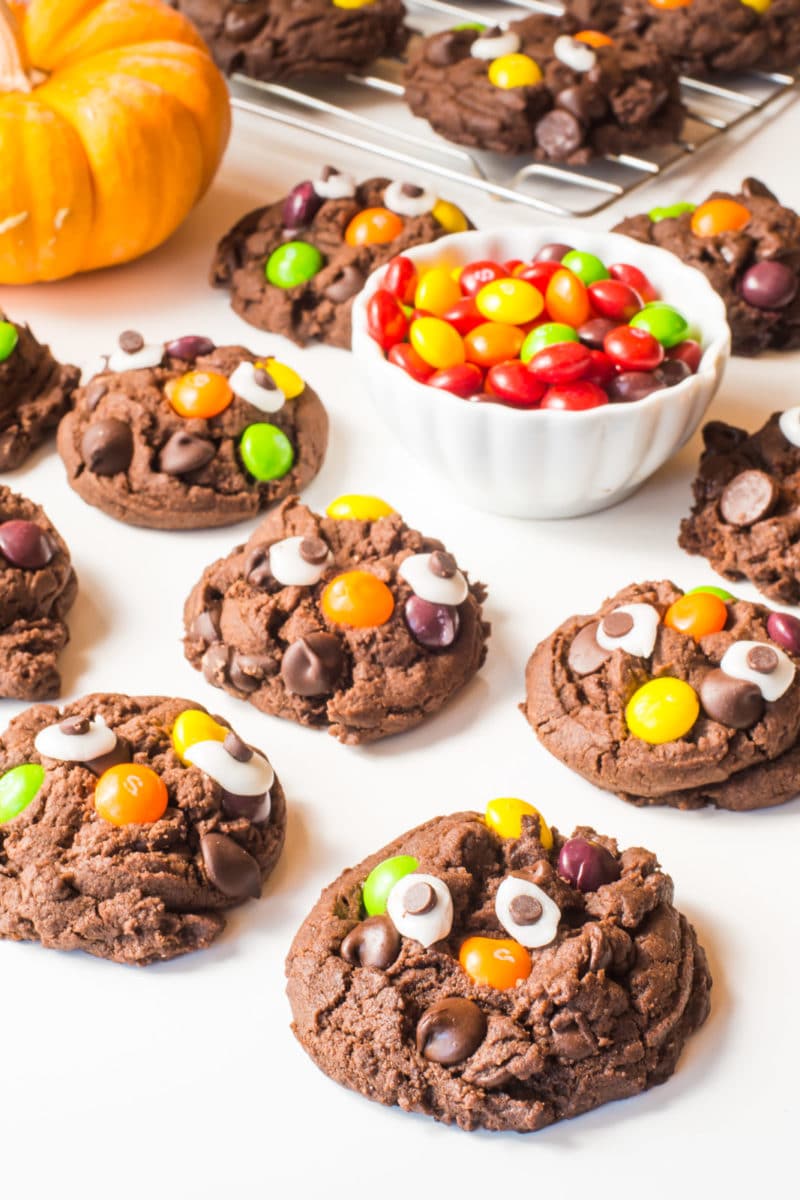 Chocolate Halloween monster cookies have candy eyes and skittles on them. They're sitting next to a bowl of skittles and a mini pumpkin.
