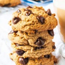 A stack of four pumpkin spiced chocolate chip cookies sit one on top of the other. The chocolate chips are shiny because the cookies are still warm. They are sitting next to a cup of hot tea with frothed almond milk. There are more cookies sitting on a wire rack in the background.