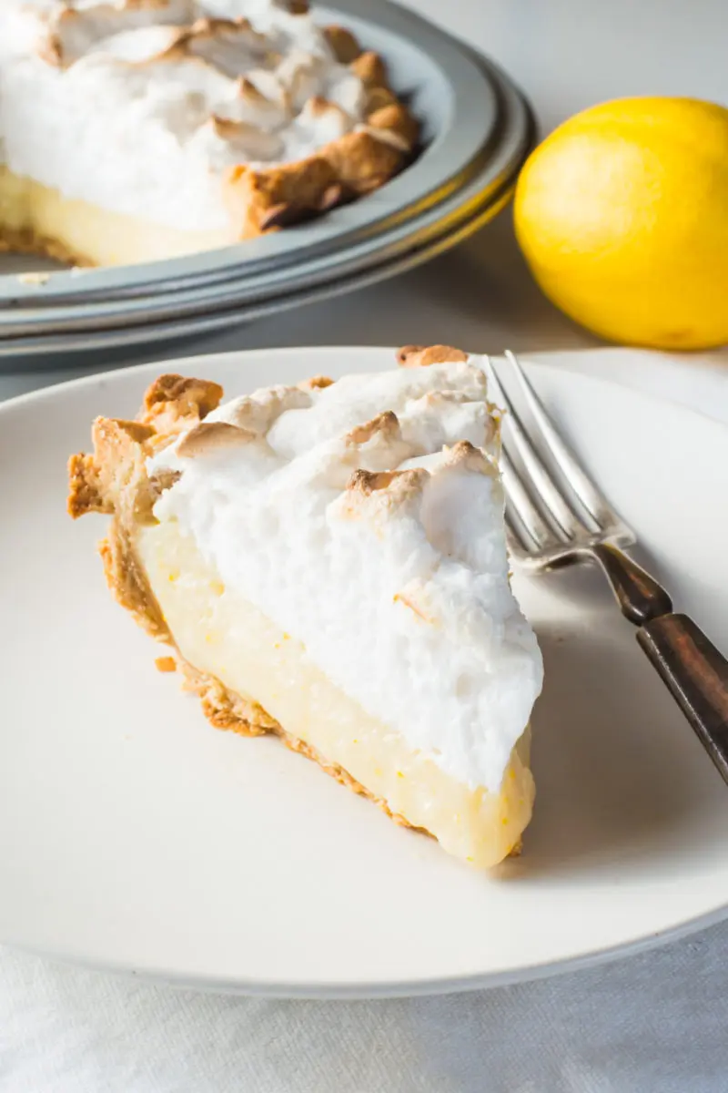A slice of lemon pie on a plate with a  fork beside it. A lemon and the rest of the pie is behind it.