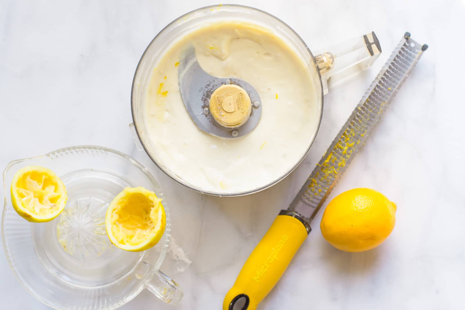 Overhead view of a food processor bowl containing meringue, a lemon, lemon zester and juicer.