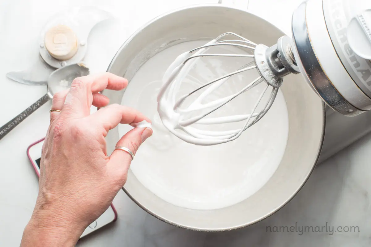 Looking down on a stand mixer with white frothy, ingredients in the bowl. A hand has some of the ingredients and is pinching it.