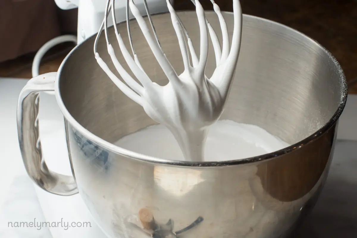 A stand mixer has vegan meringue inside that is falling from a whisk attachment into the bowl.