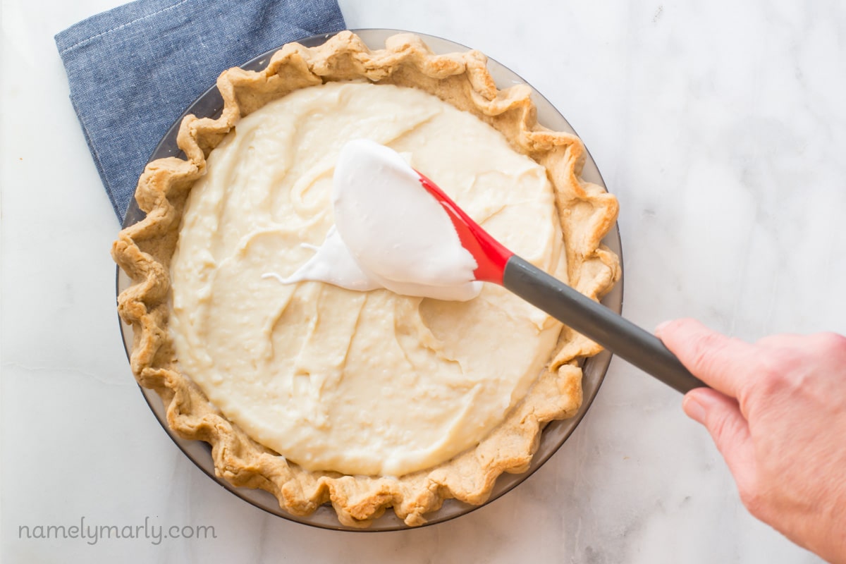A hand holds a spatula, spreading cream filing across a pie.