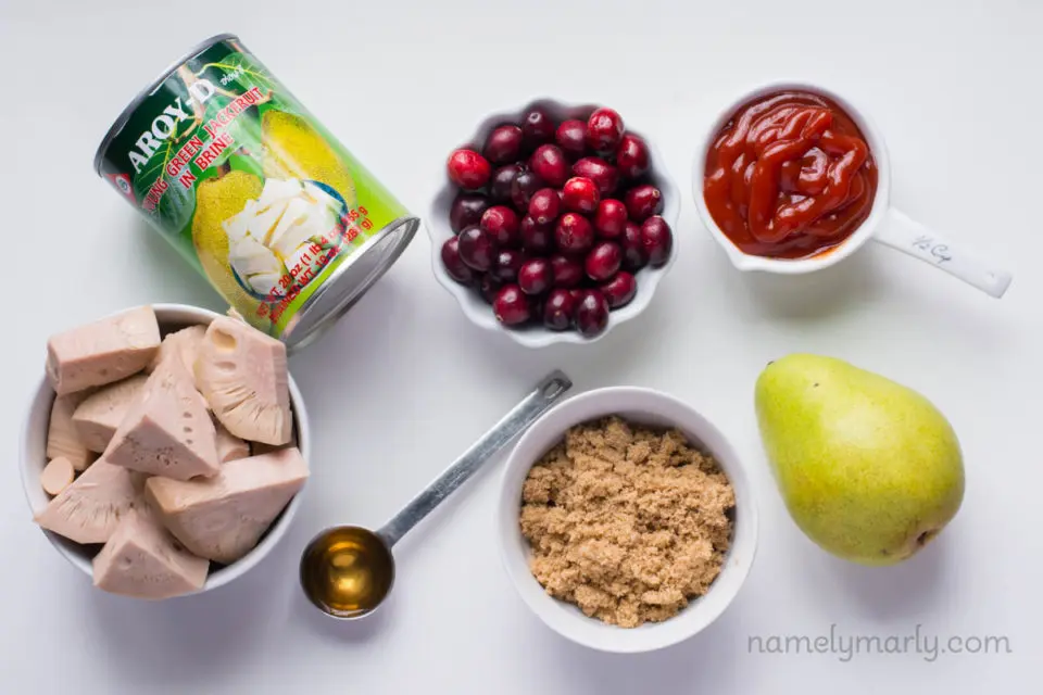 The ingredients to make BBQ jackfruit on a white counter top, including canned jackfruit, brown sugar, a pear, cranberries, and more.