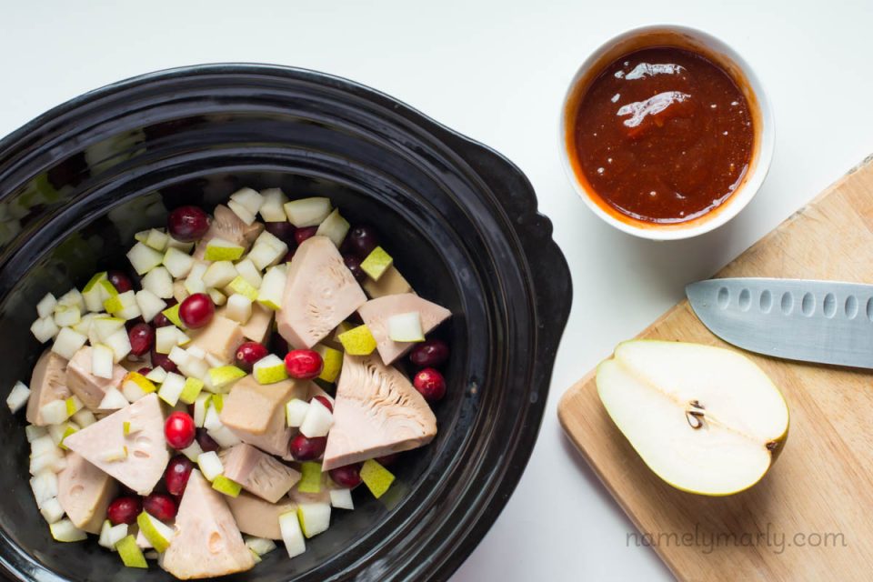 A slow cooker is filed with canned jackfruit wedges, and other ingredients. Next to it is a bowl of BBQ sauce and a halved pear.