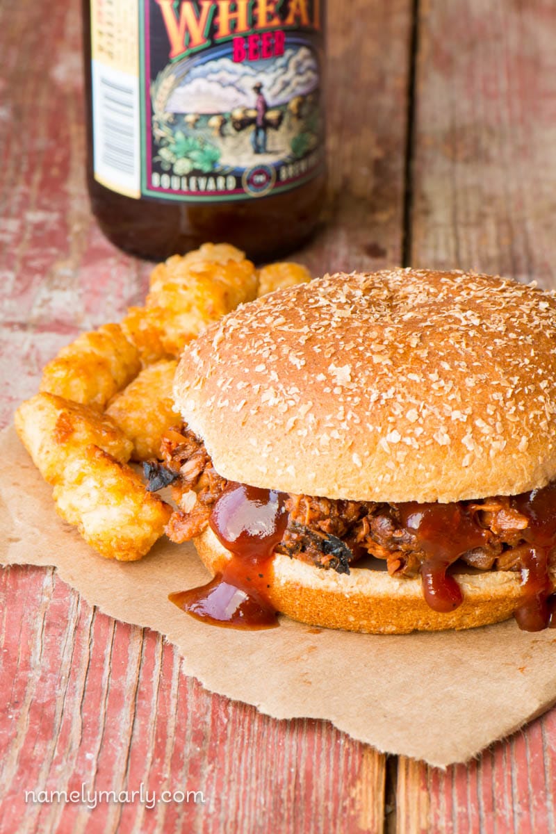 Some Slow Cooker BBQ Pulled Jackfruit sits between two hamburgers buns with BBQ sauce drizzled over the edges. There are tater tots and a bottle of beer behind it.