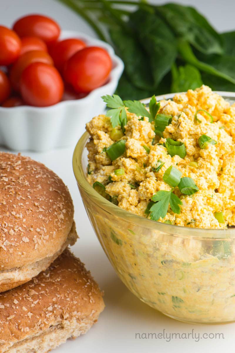 A bowl holds a batch of vegan egg salad. It sits besides stacked hamburger buns, a bowl of cherry tomatoes, and greens.