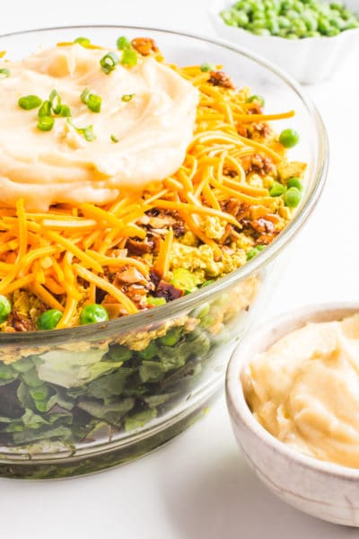 A close-up of Vegan Seven Layer Salad in a large bowl with frozen peas behind it.