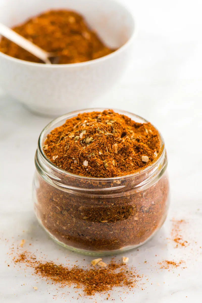 A small glass jar is full of homemade taco seasoning sits in front of a bowl with more of the seasoning.