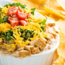 A dish full of vegan taco dip is topped with vegan cheddar shreds and sliced tomatoes. There are tortilla chips behind it.