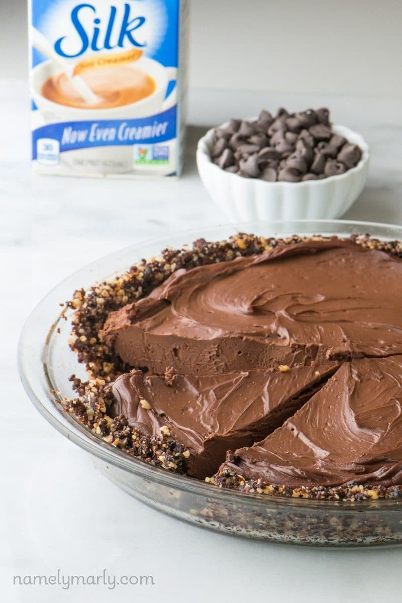 A pie pan holds vegan chocolate pie, with one slice cut out. A carton of plant-based creamer is behind it and a bowl of chocolate chips.