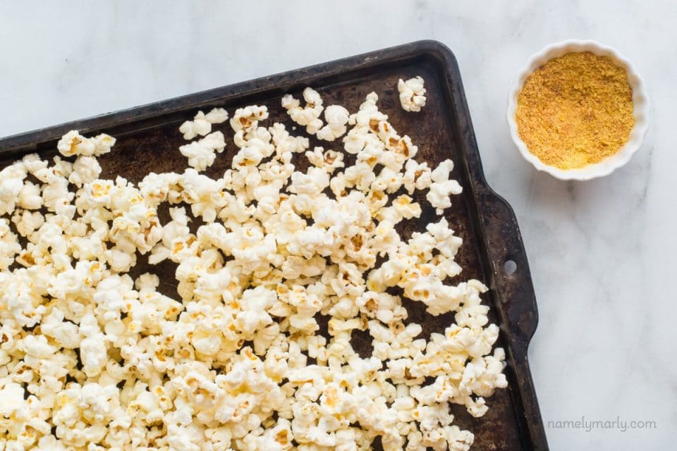 A baking sheet is full of freshly popped popcorn with a bowl of nutritional yeast flakes beside it.