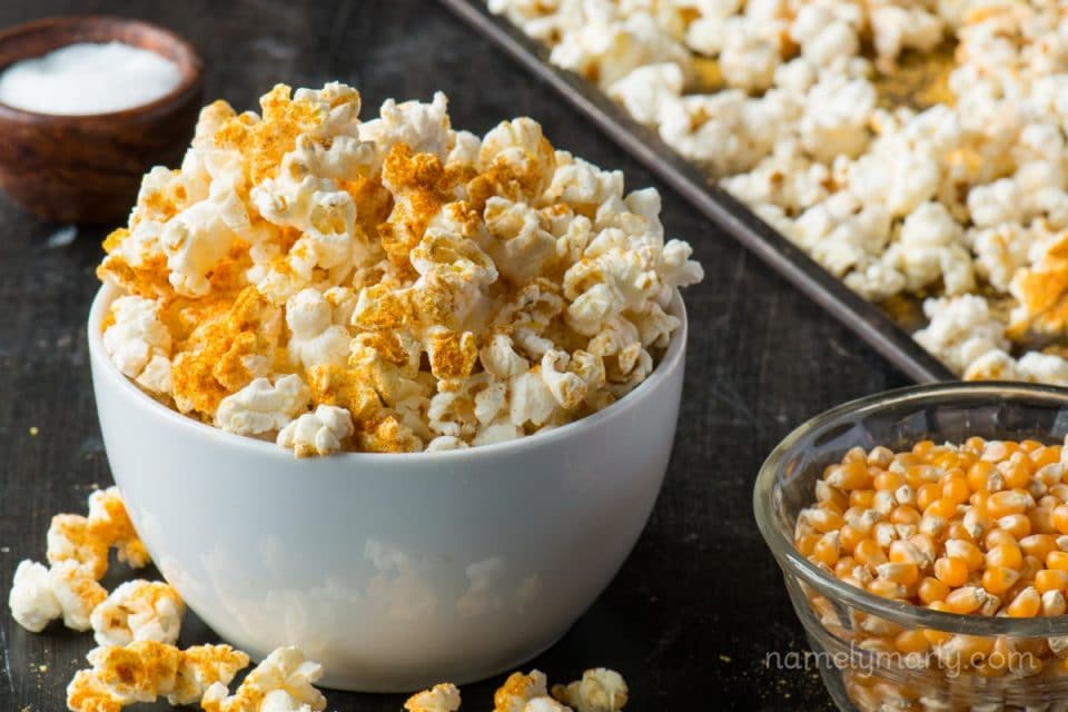 A bowl of popcorn sits on a baking sheet with lots of popped popcorn around it, a bowl of salt and unpopped popcorn beside it.