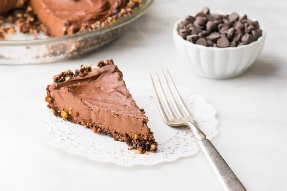 A slice of vegan chocolate pie on a doily next to a fork with a bowl of chocolate chips and the rest of the pie behind it.