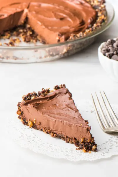 A slice of vegan chocolate pie on a doily next to a fork. The rest of the pie is behind it next to a bowl of chocolate chips.
