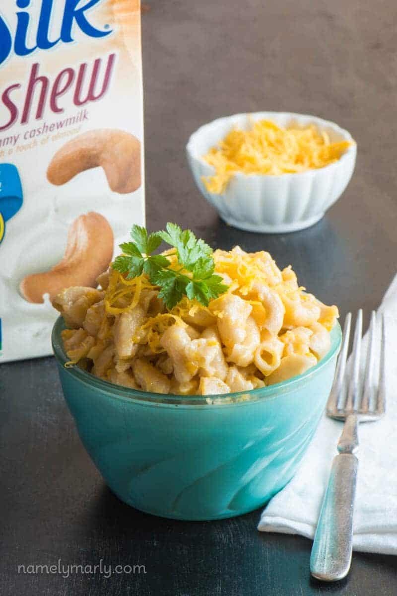 A bowl of vegan mac and cheese is topped with fresh parsley sprigs. A fork is beside it. A carton of plant-based milk and a bowl of vegan cheddar shreds is behind it.