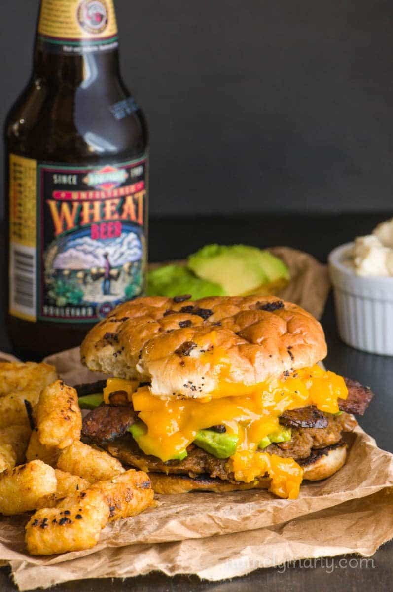 A veggie burger sits on brown paper with tater tots beside it. A bottle of beer is behind it with more avocado slices and vegan mayo.