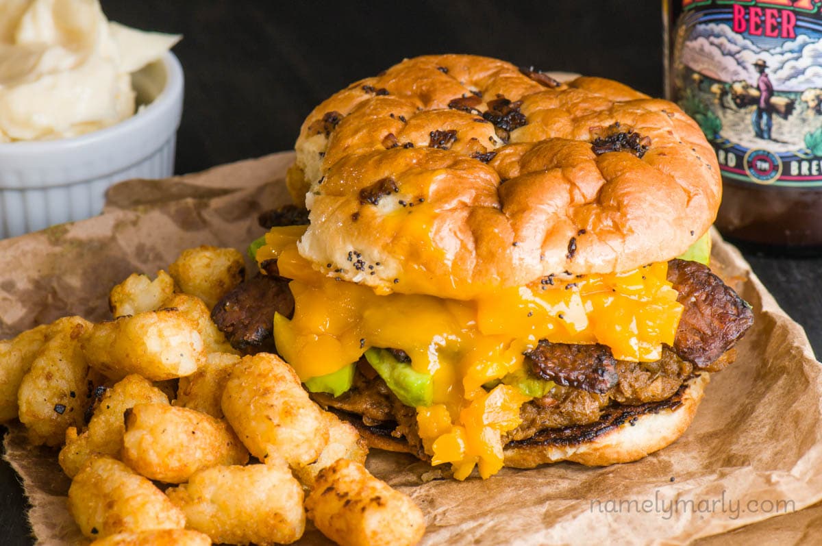 A veggie burger sits on a brown bag with tater tots beside it. A bottle of beer is on one side of the burger and a bowl of mayo is on the other side.