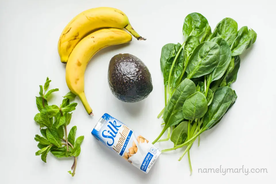 Ingredients to make a vegan shamrock shake on a white counter. These include an avocado, plant-based milk, fresh spinach, mint sprigs, and a couple of bananas.