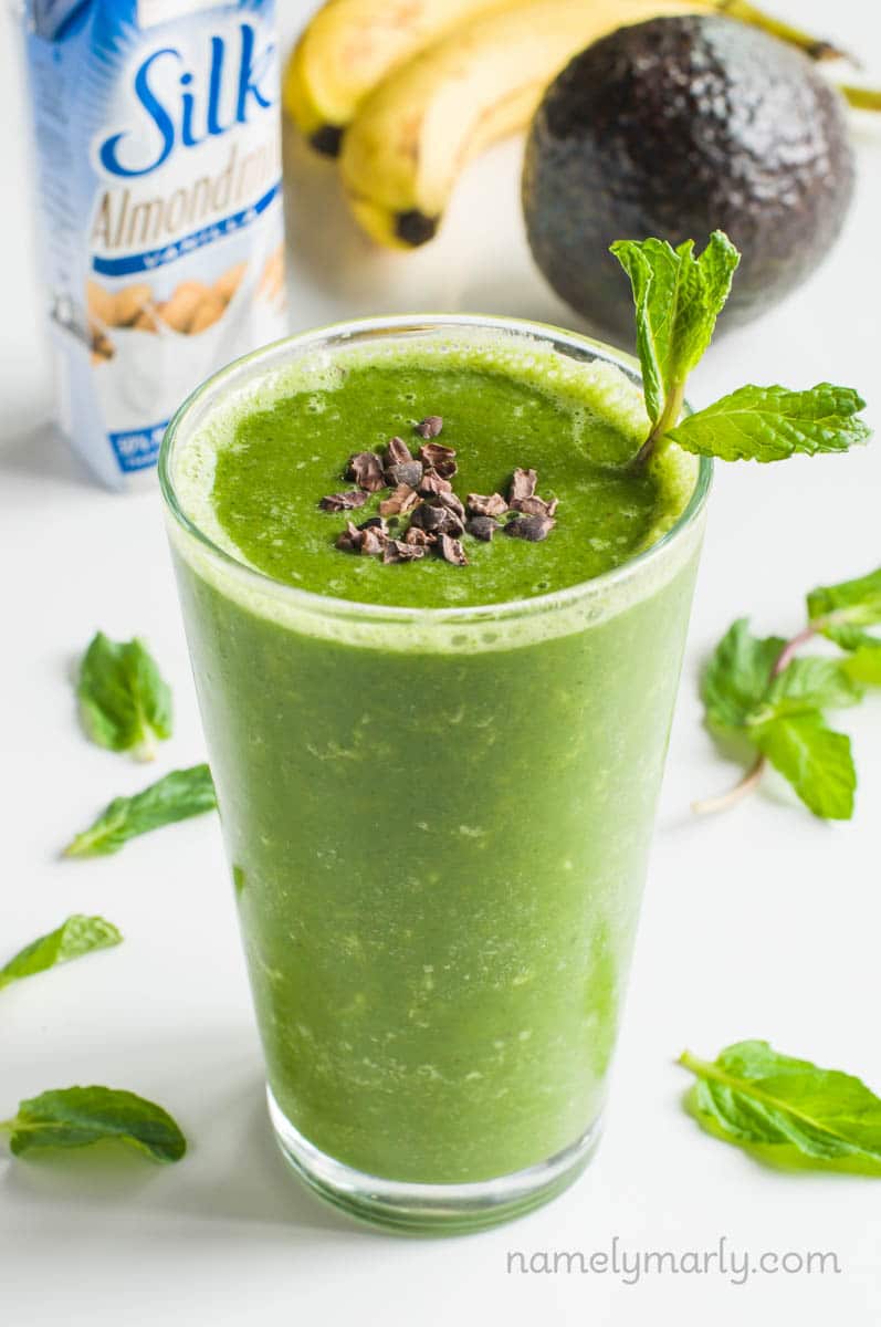 A green smoothie in a tall glass has cacao nibs and mint sprigs in it. There's mint sprigs, an avocado, banana, and a carton of plant-based milk behind it.