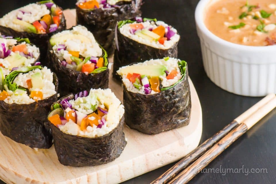 Slices of vegan sushi sit on a cutting board with chopsticks sitting in front, a bowl of dipping sauce sits nearby.