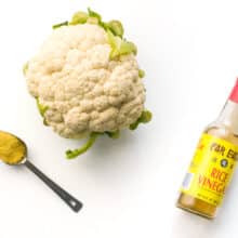 A head of cauliflower sites next to a spoon full of nutritional yeast flakes and rice vinegar.