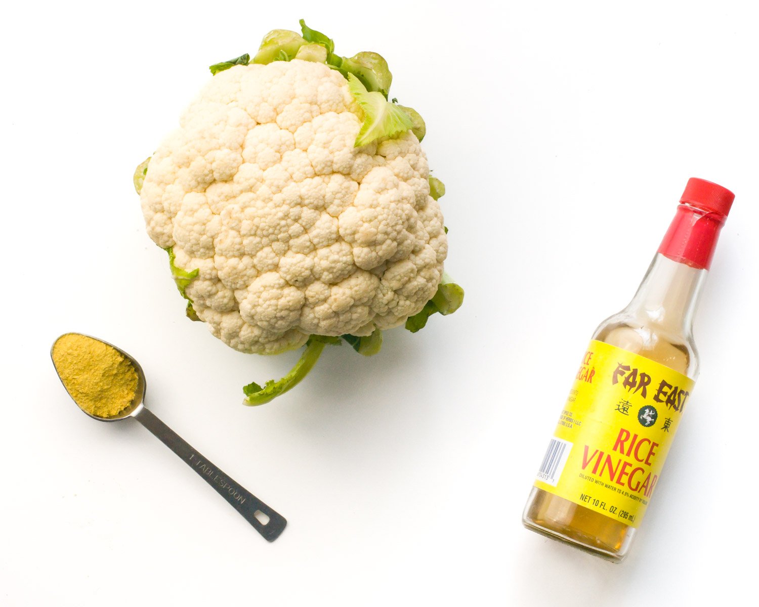A head of cauliflower sites next to a spoon full of nutritional yeast flakes and rice vinegar.
