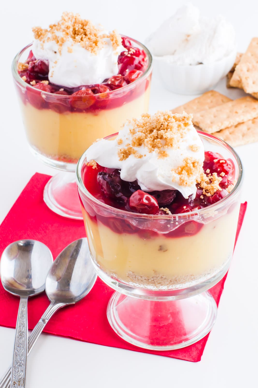 Two serving dishes full of vegan cherry vanilla parfaits are topped with whipped cream. Spoons sit beside them and graham crackers and more whipped cream is behind them.
