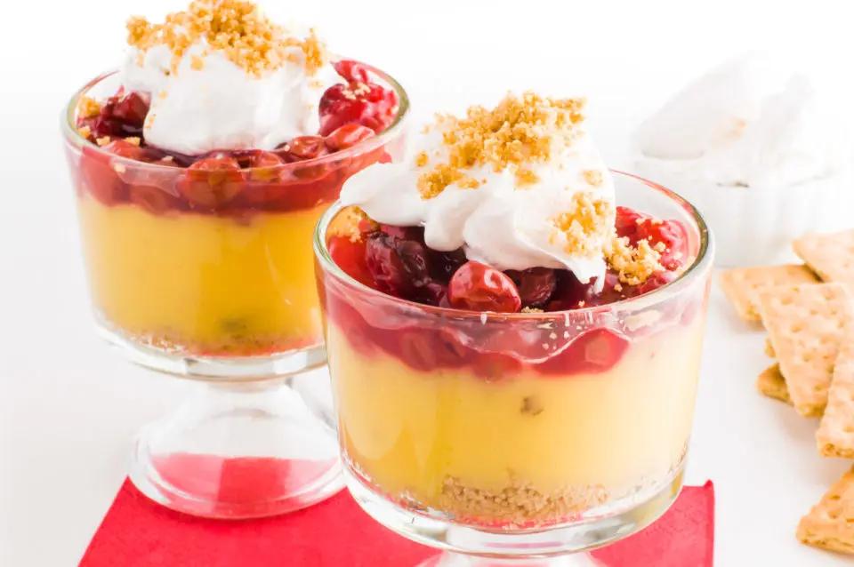 Two vegan parfaits topped with cherries, whipped cream, and graham crackers. There are more graham crackers and a bowl of whipped cream behind it.