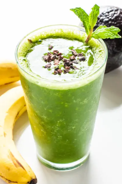 A green smoothie in a tall glass has cacao nibs and mint sprigs in it. There are bananas next to it and an avocado behind it.