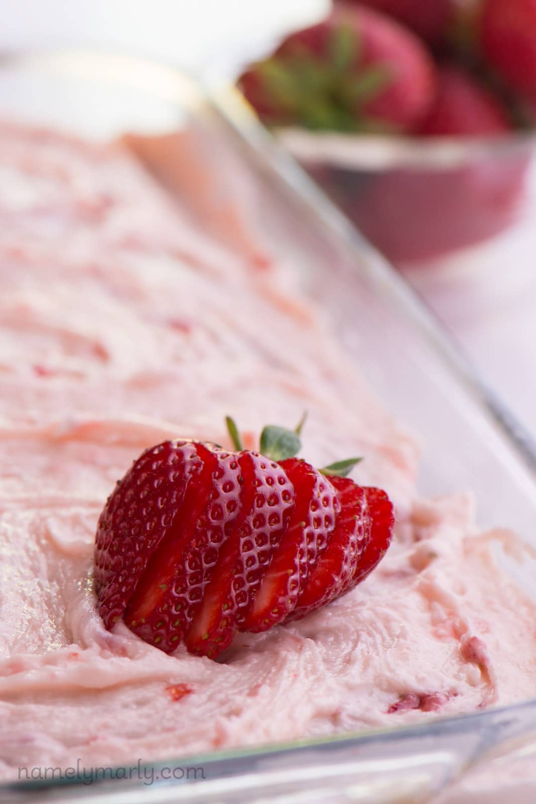 A sliced strawberry sits on top of a 9X13 strawberry cake. A bowl of fresh strawberries is in the background.