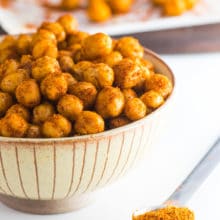 A bowl is full of roasted chickpeas with a teaspoon of seasoning beside it.