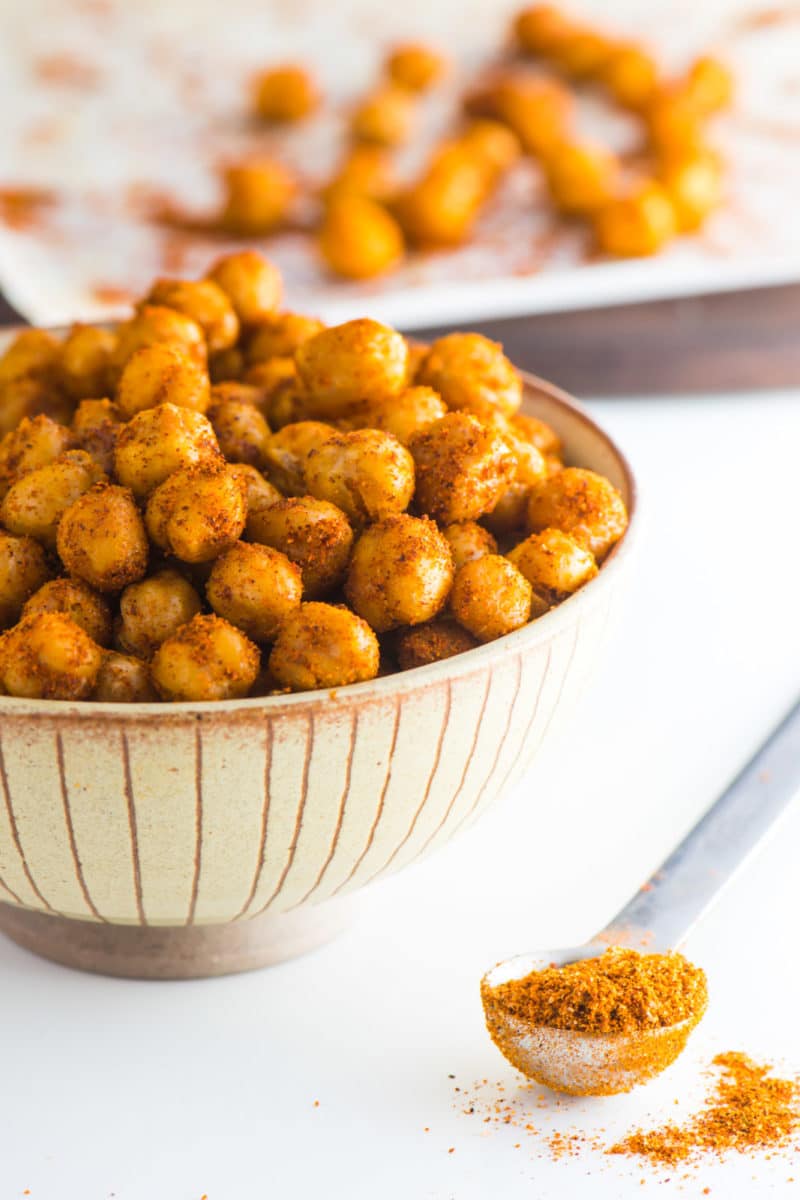 Roasted chickpeas are in a bowl with a measuring spoon of spices beside it. There are more roasted chickpeas on a pan in the background.