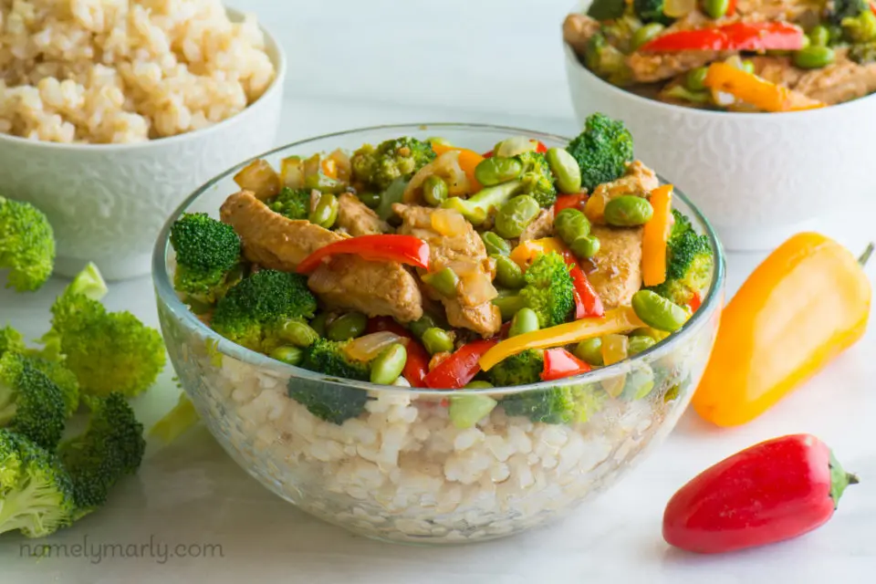 A bowl of brown rice topped with a vegan teriyaki mixture. It's surrounded by bowls of brown rice, more teriyaki mix, and steamed broccoli and mini bell peppers.
