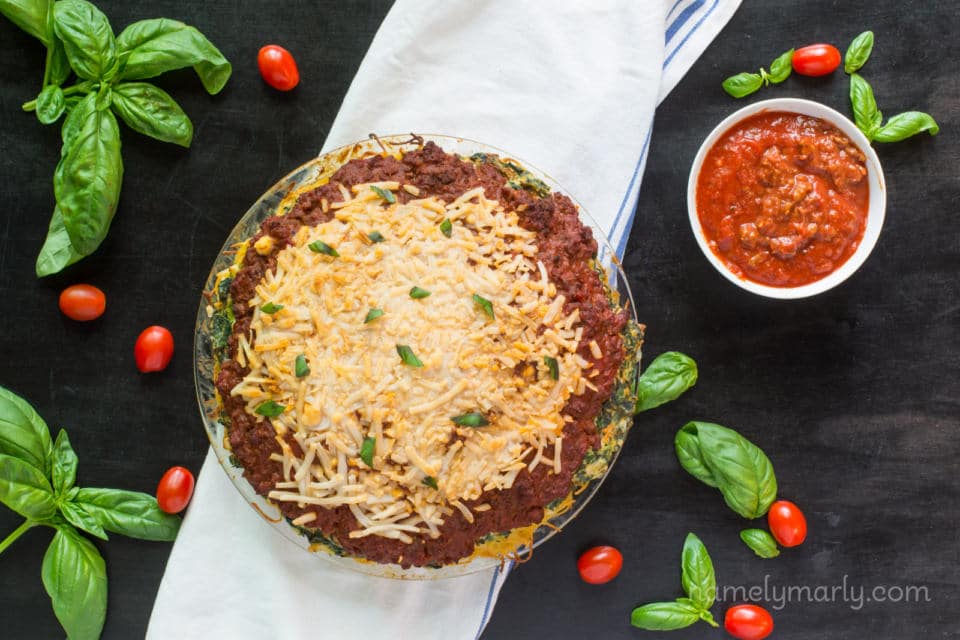 We look down at a pie pan full of vegan spaghetti pie, freshly pulled from the oven and sitting on a white kitchen towel. There's fresh basil and cherry tomatoes arranged on the dark counter and a bowl of additional sauce.