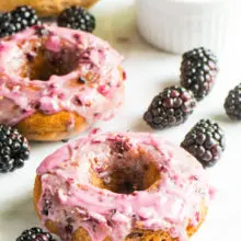 Two vegan blackberry donuts sit next to blackberries. A bowl of blackberry glaze and a stack of unglazed donuts sits behind it.